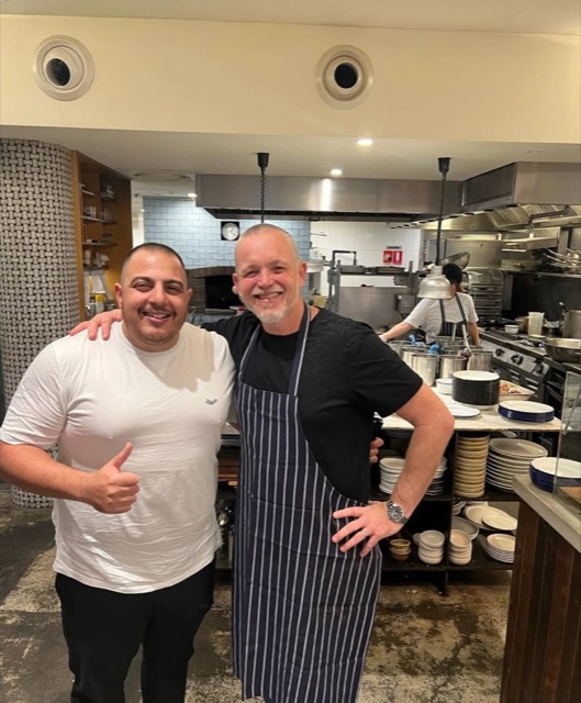 Paul with a chef in a restaurant kitchen with a wood-fired oven